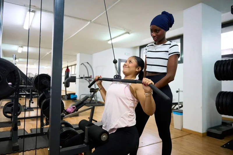 Two individuals in a gym one using an exercise machine as the other assists