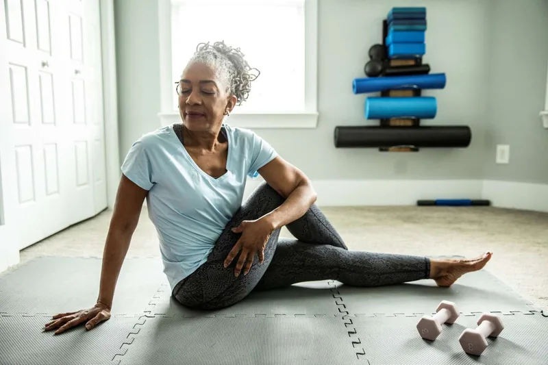 A woman seated on an exercise mat in a stretching pose with fitness equipment in the background