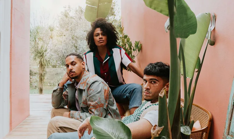 Three stylish young people sit on a rattan couch against a pink wall, surrounded by lush greenery. They all confidently look at the camera. They exude individuality and determination.