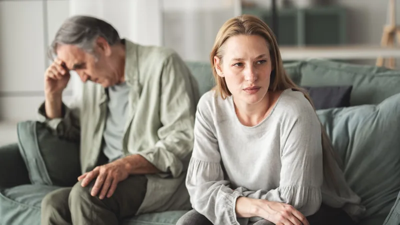 A woman sits on a couch looking away while an older man sits behind her with a contemplative pose