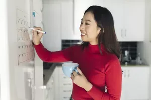 Smiling woman with coffee cup writing on calendar in kitchen