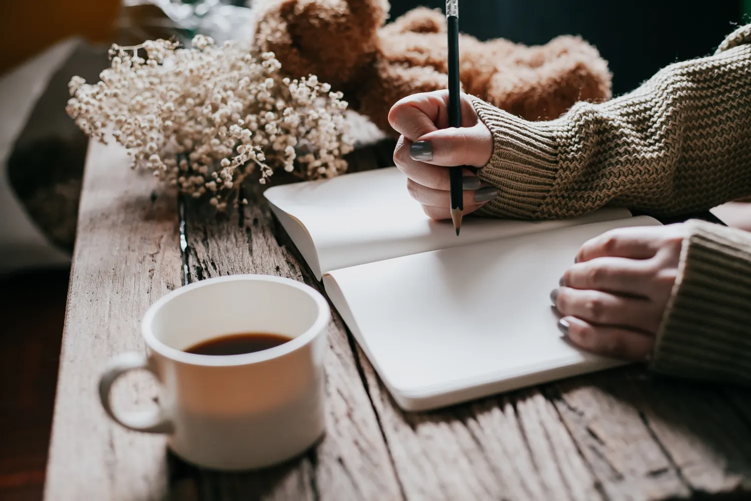 A person writing in a notebook on a wooden table next to a cup of coffee and a floral arrangement