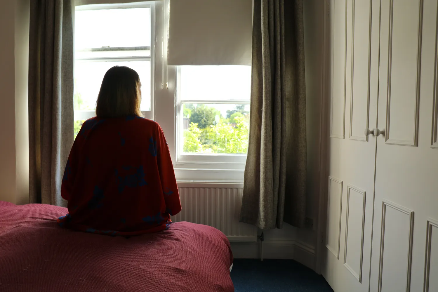 Woman wearing maroon robes, sitting on pink sheets staring outside into a window exposing trees