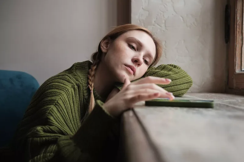 Upset sad millennial girl sitting by window looking at mobile phone waiting call from boyfriend