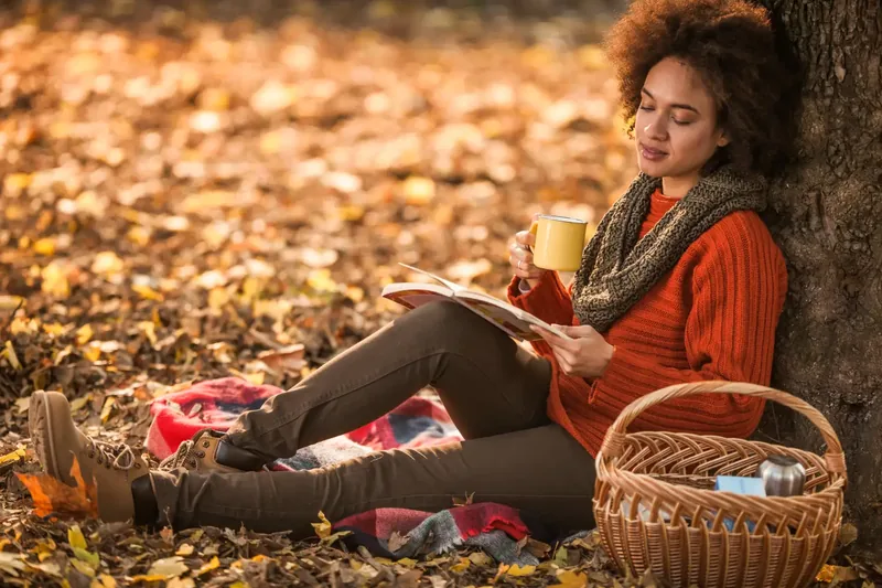 Person sitting under a tree in an autumnal setting with a book and a mug a basket with items nearby