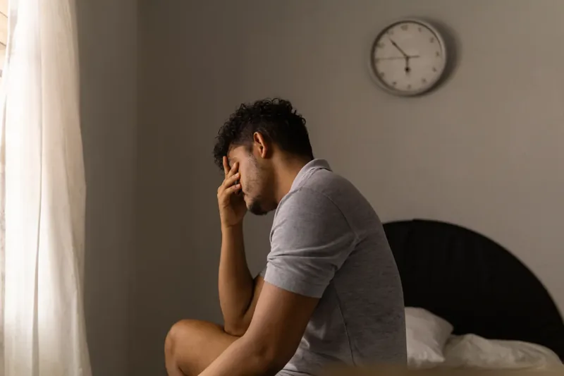 A person sitting on a bed resting their face in their hand with a wall clock in the background