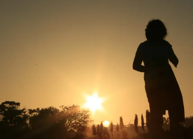 A person stands on a mountain peak, arms outstretched, looking out over a vast, misty landscape with rolling hills and a winding road.