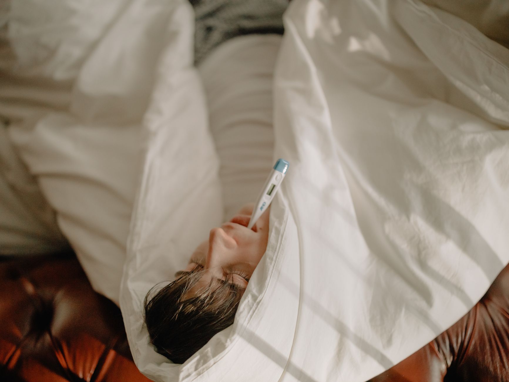 A man lying in bed with a thermometer in his mouth, wrapped in cozy bedding, indicating illness or fever.