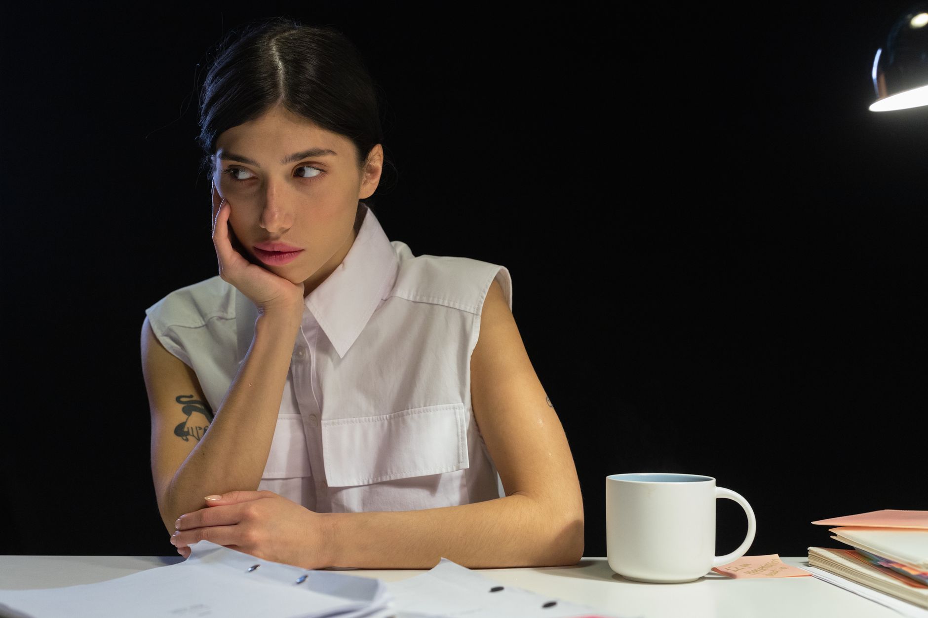 A thoughtful woman sits at a desk with papers and a mug, illuminated by a lamp.