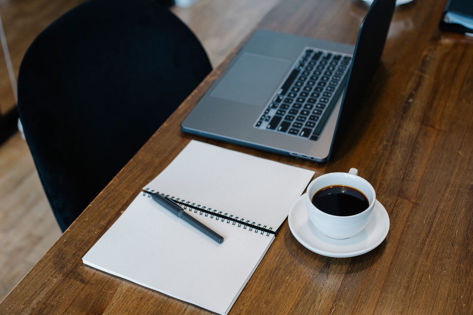 High angle of opened laptop and spiral notebook with blank pages placed on wooden table with cup of coffee and pen during workday