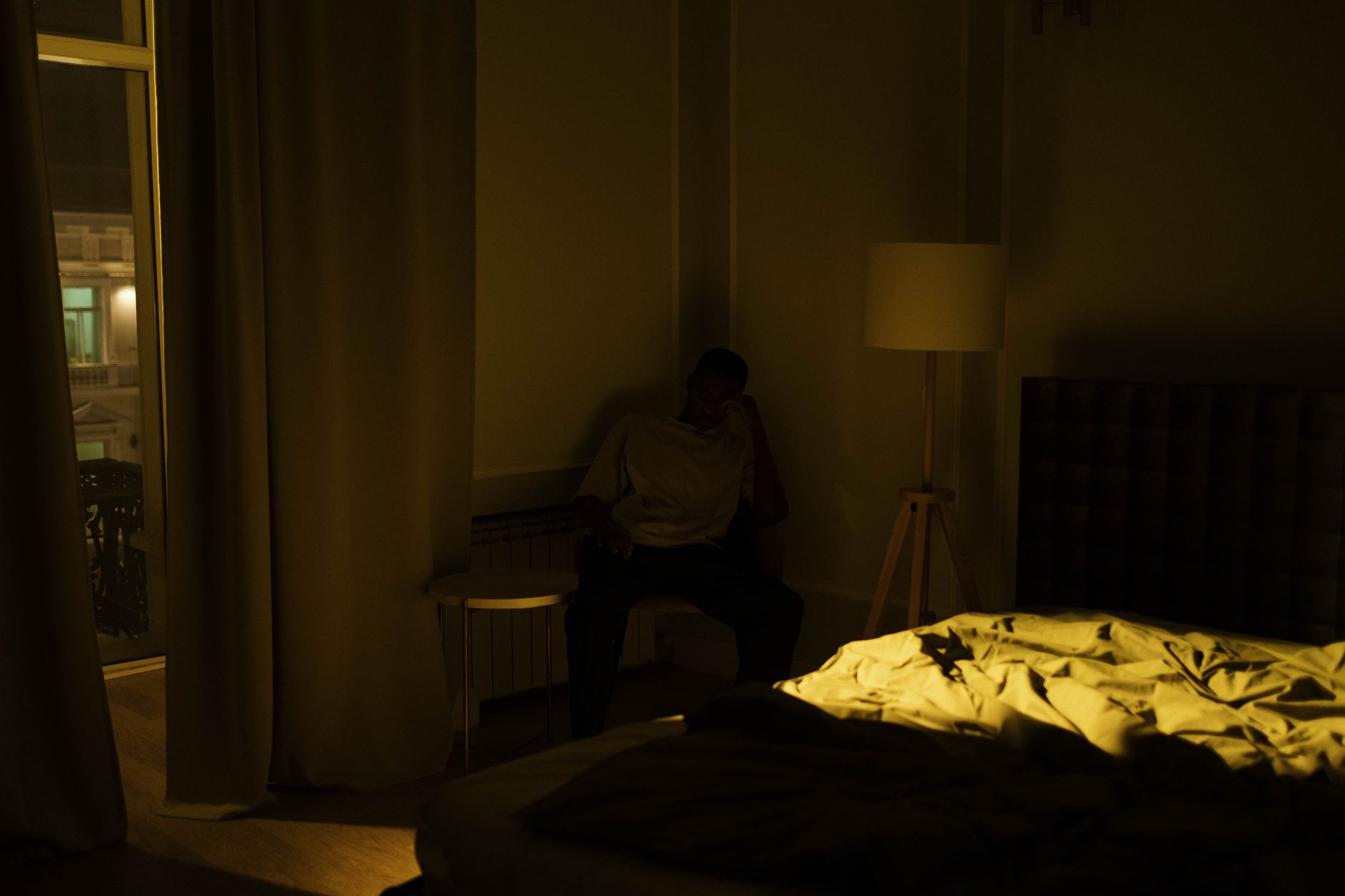 An adult man experiencing insomnia, sitting in a dimly lit bedroom corner at night.