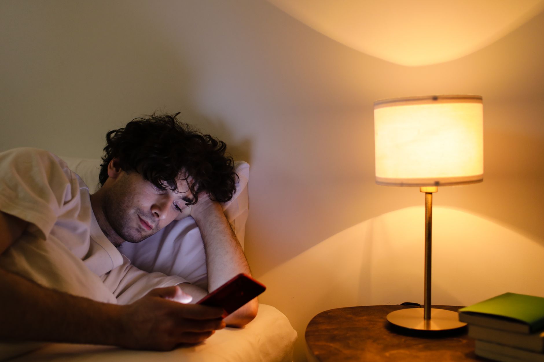 A man lying in bed using a smartphone with a lamp providing warm ambient light.