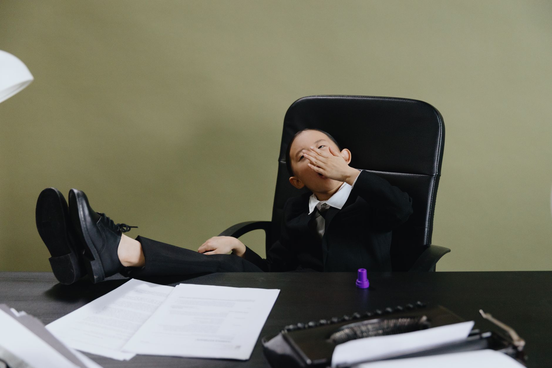 A young child in a black suit yawns while sitting at an office desk, suggesting fatigue or boredom.
