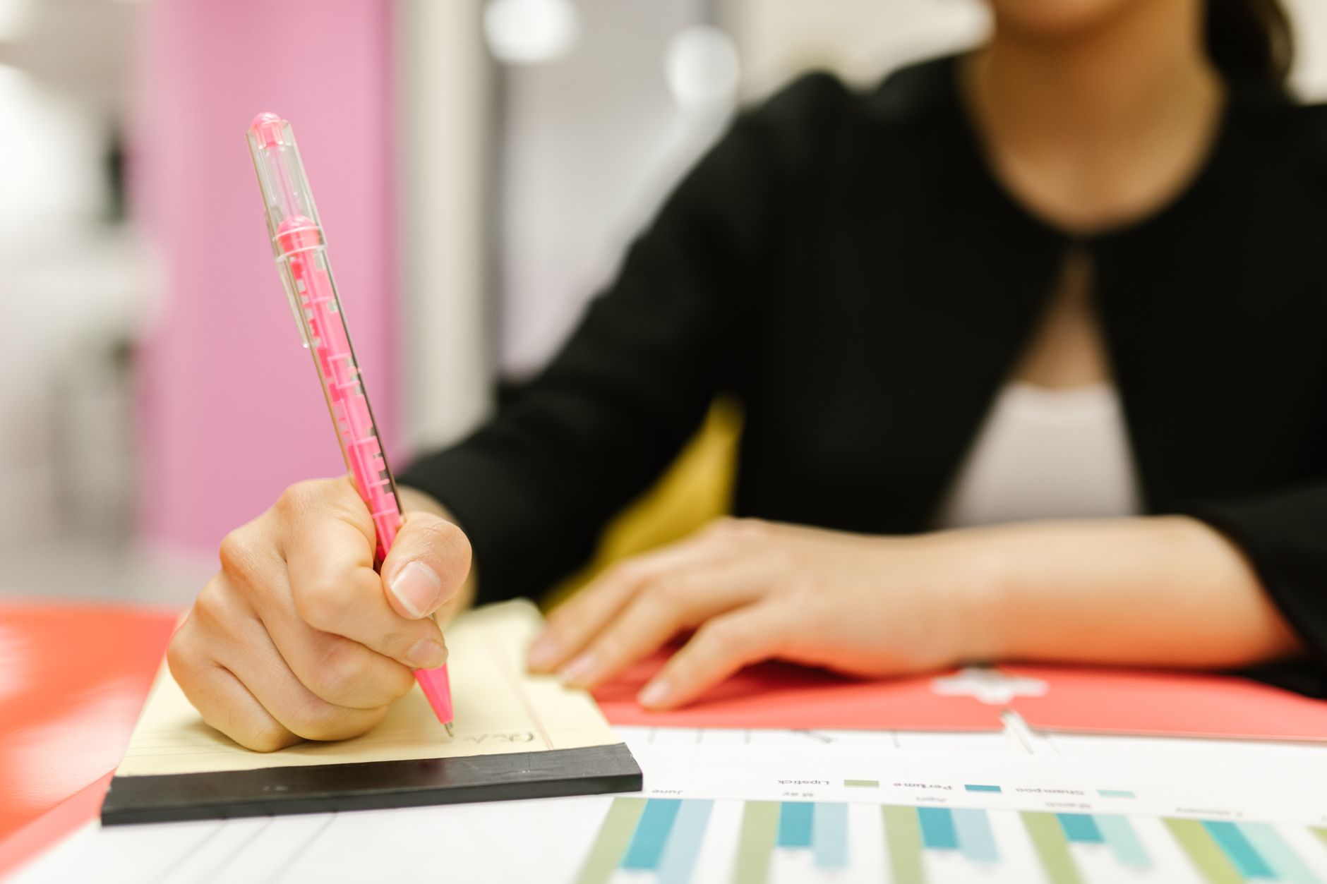 A woman writing notes on a notepad with colorful charts in a modern office setting.