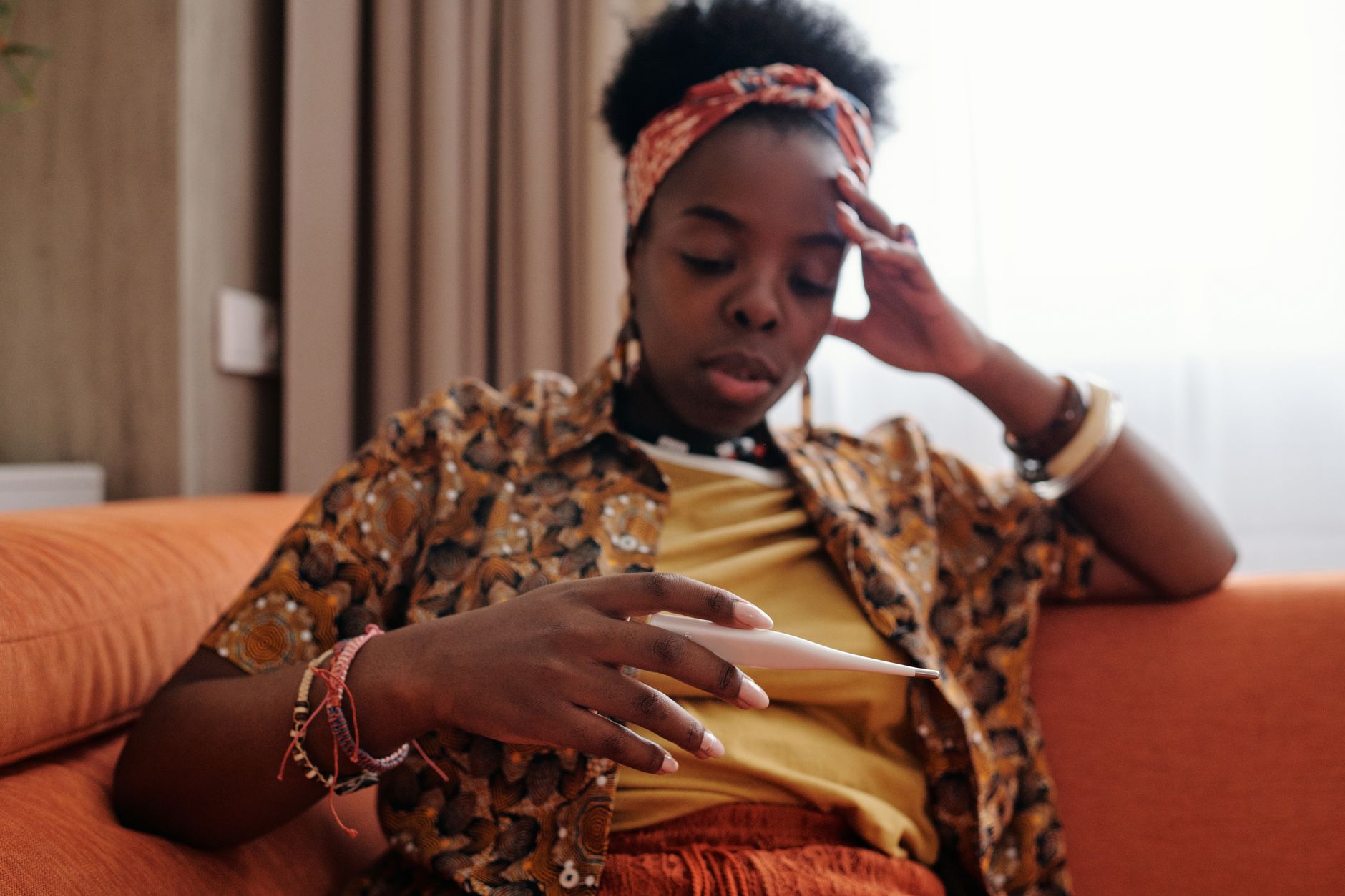 A woman looks at a thermometer with concern, sitting indoors on a couch.