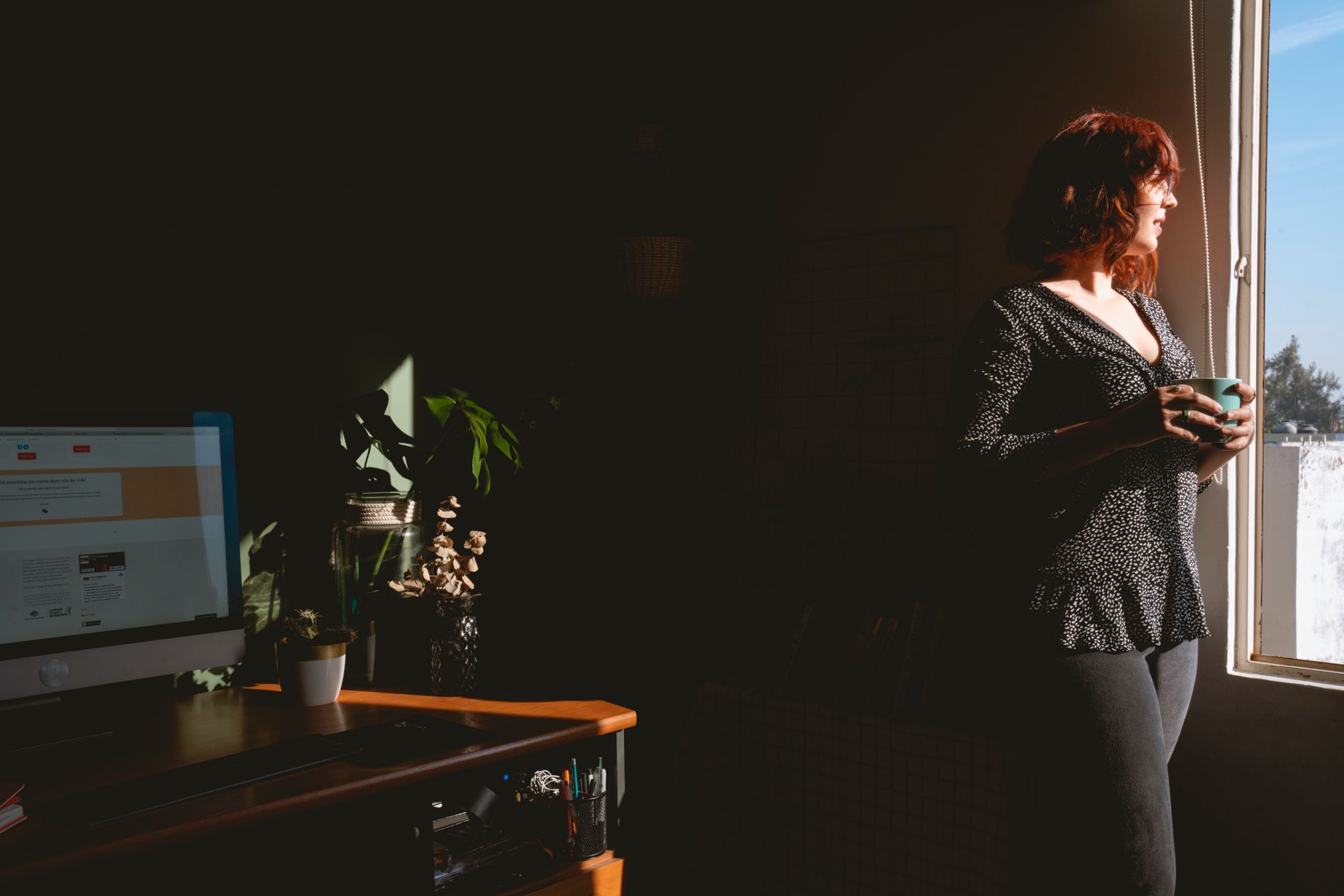 A woman stands in a home office holding a mug, bathed in natural light from a window.