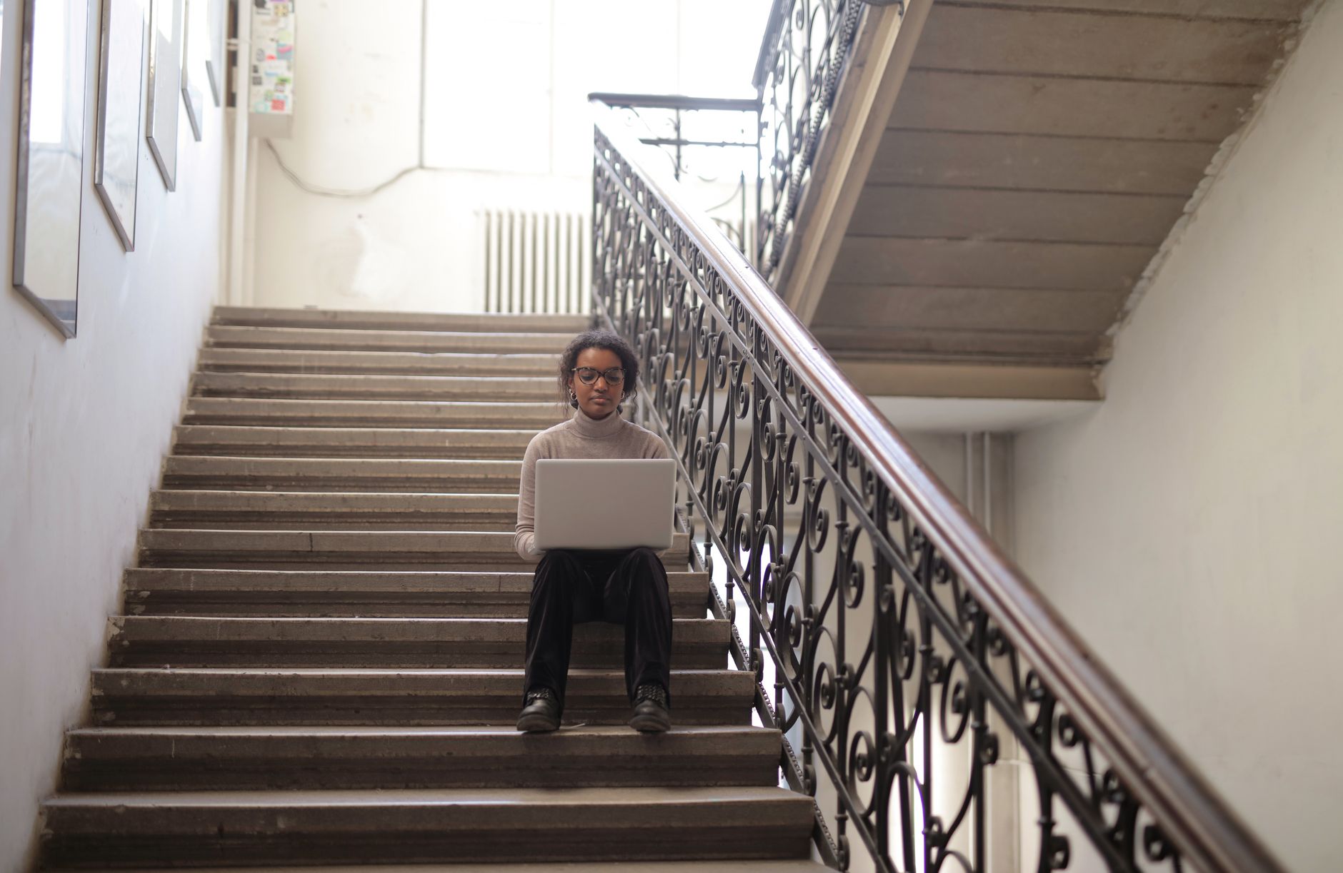 A woman sits with a laptop on a staircase, working in a bright indoor setting.