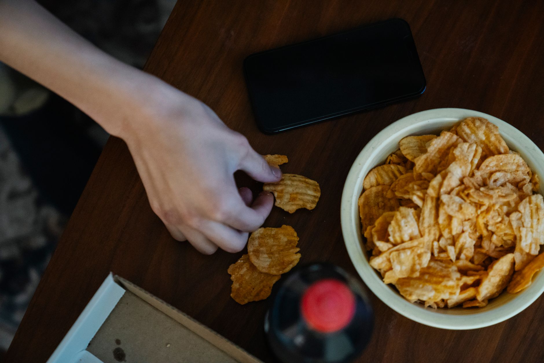 From above of crop anonymous person with crispy potato chips at table against bottle of refreshing drink and smartphone
