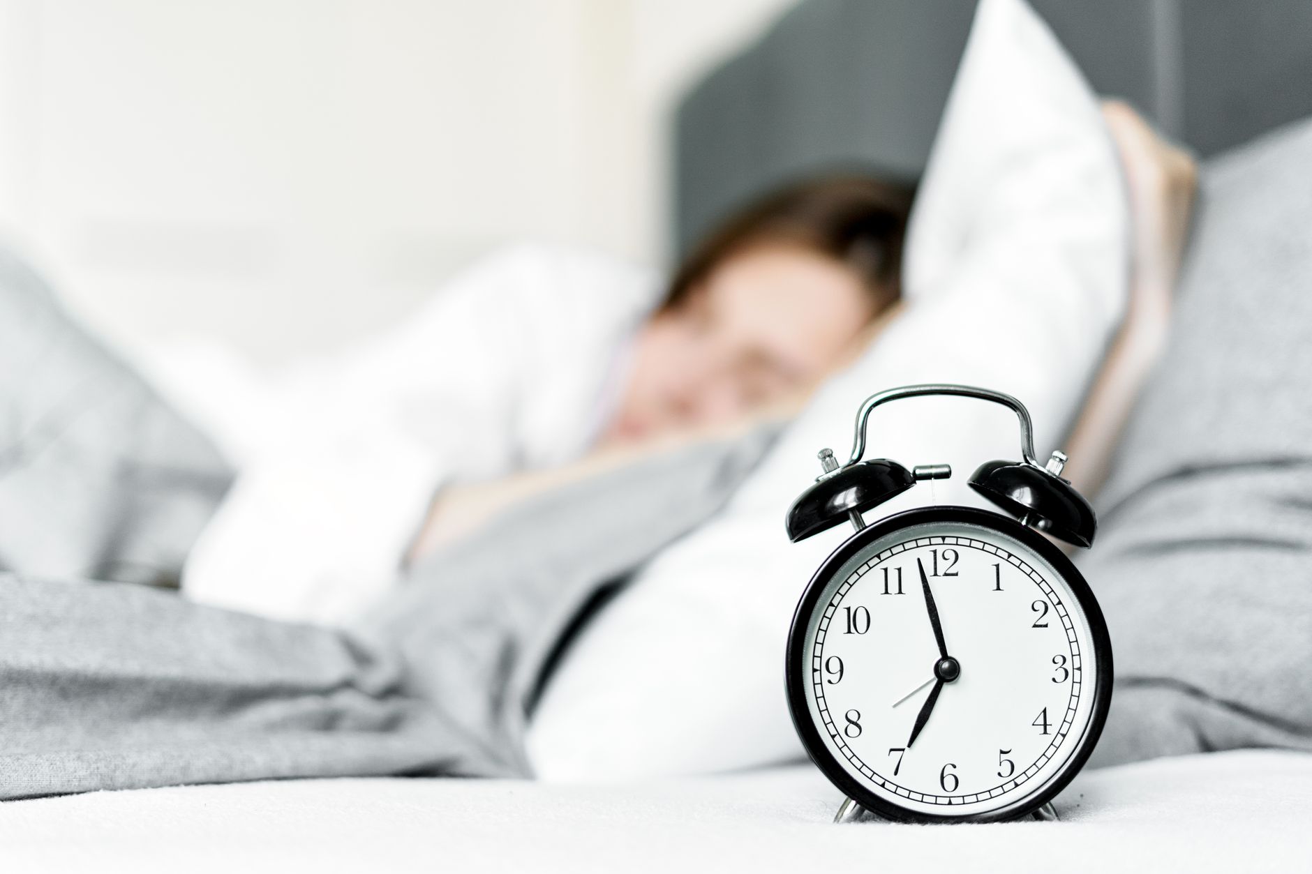 A retro alarm clock sits by a sleeping woman in a cozy bedroom setting.