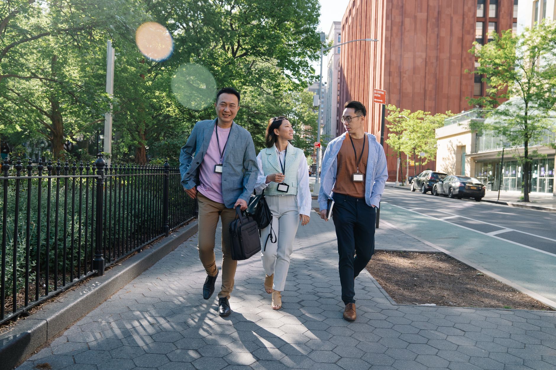 Three adults walking on a city sidewalk during a sunny day break, wearing business attire and ID badges.