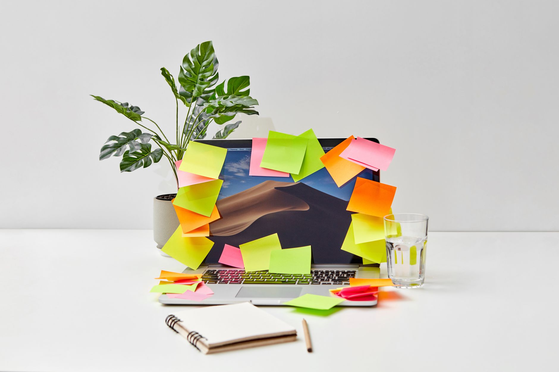 A cluttered office desk featuring a laptop covered with colorful sticky notes beside a glass of water.