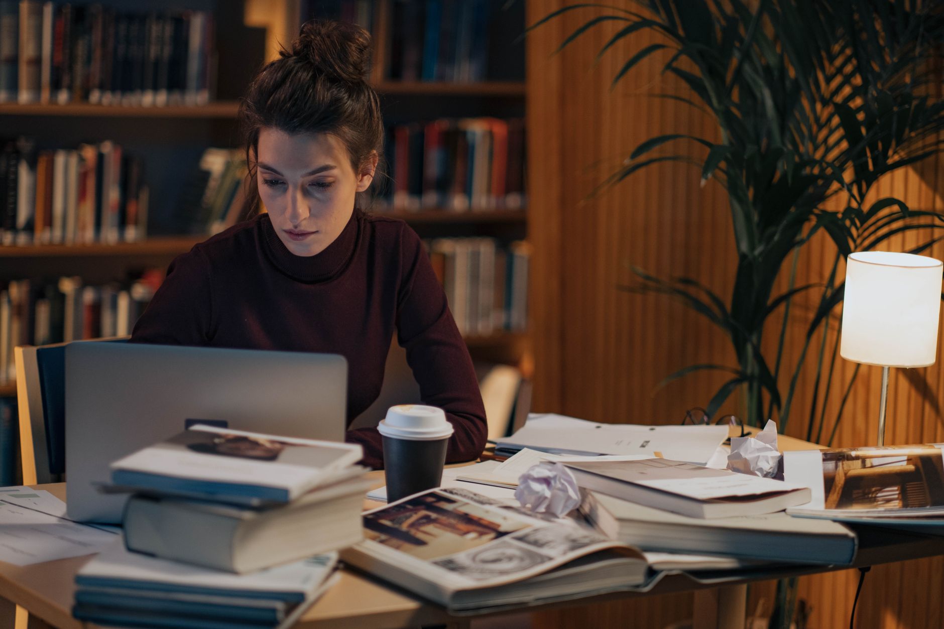 Woman working late at a desk filled with books, laptop, and coffee in a cozy library setting.