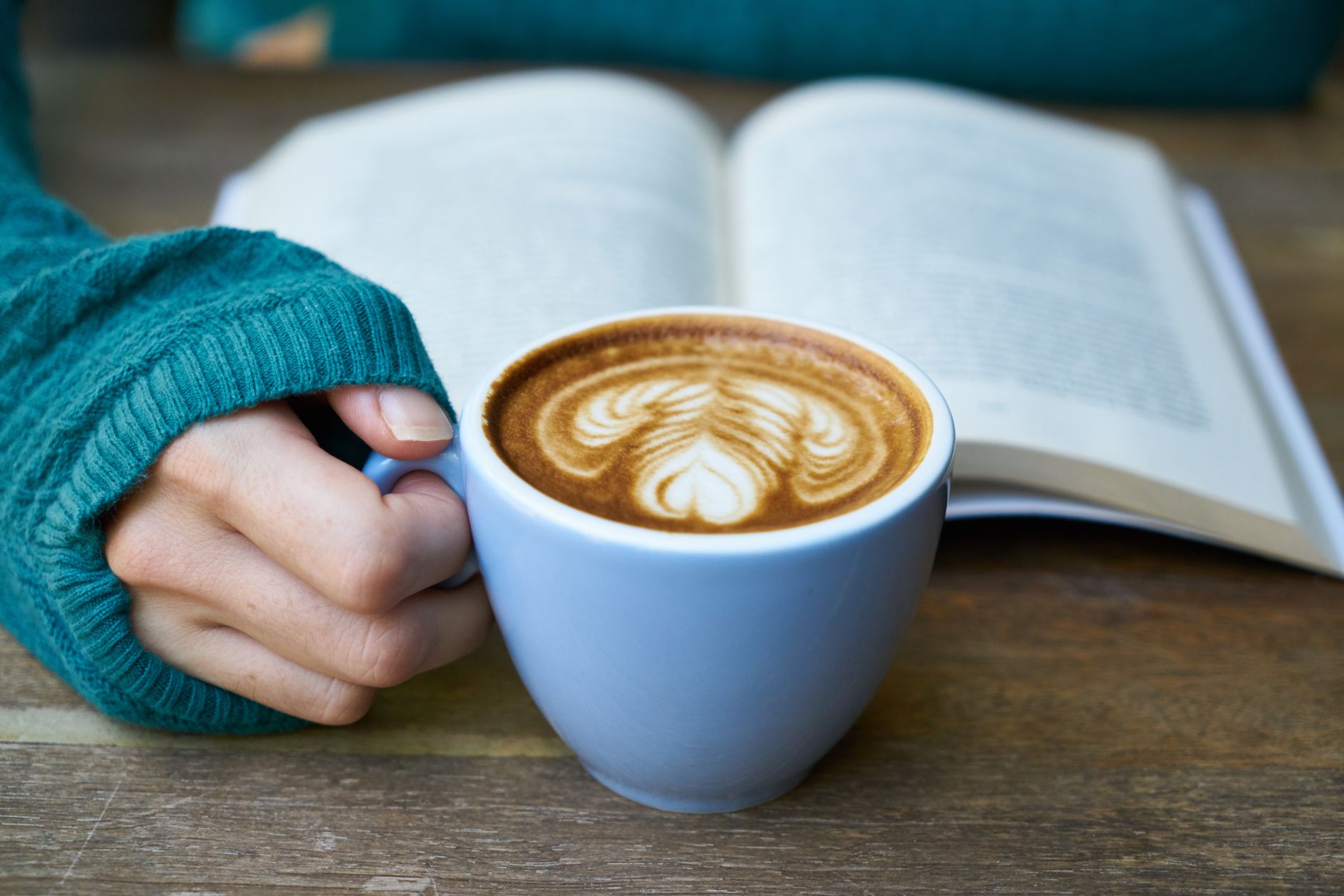 A close-up of a hand holding a latte art cup on a wooden table with an open book in the background, ideal for cozy and relaxing themes.