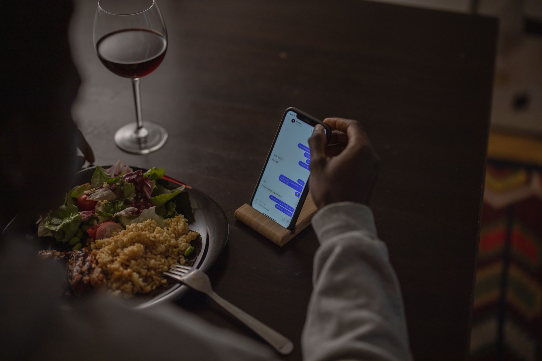 Person using smartphone at dining table with salad, grains, and red wine.