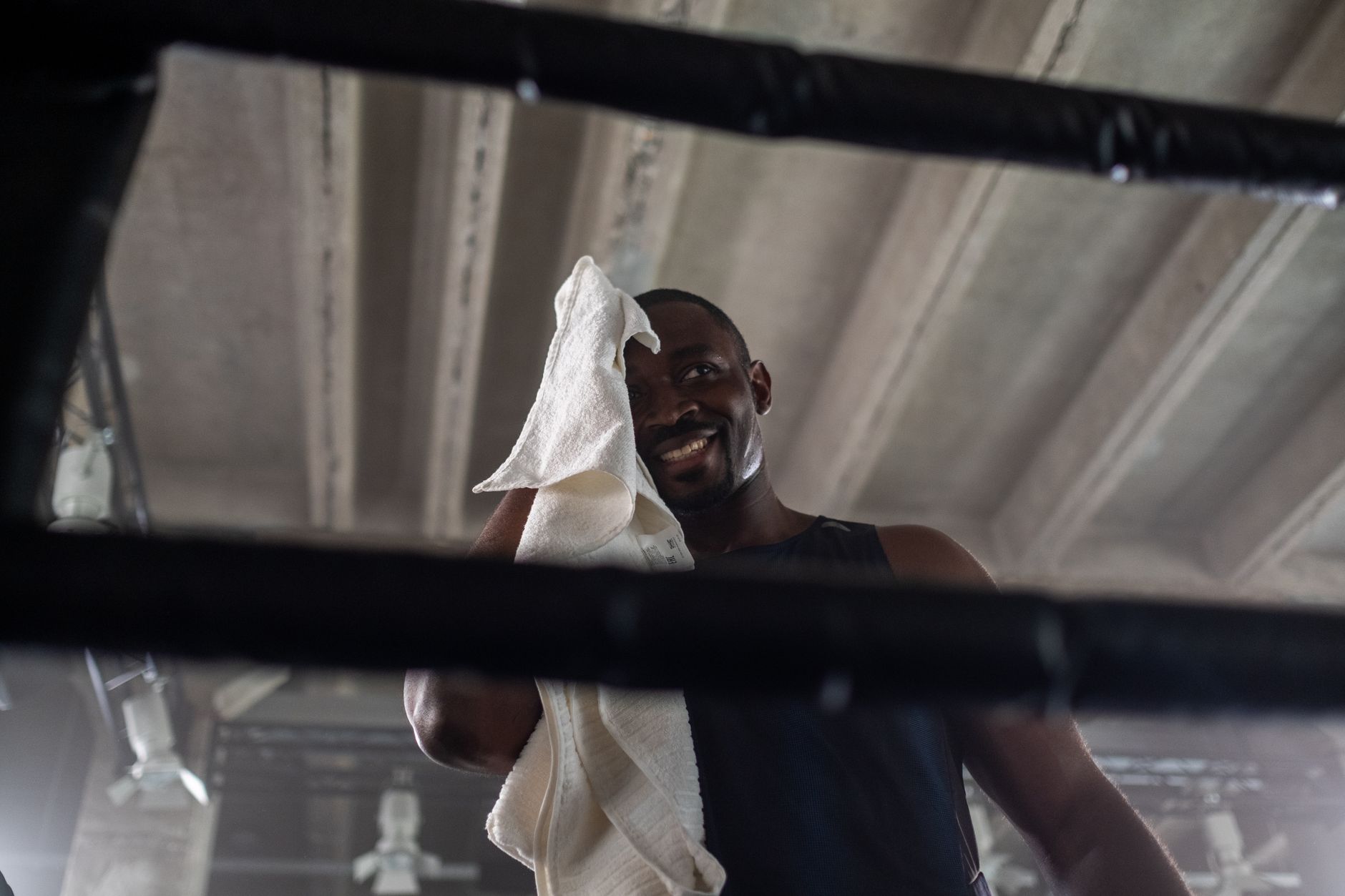 A smiling boxer wipes sweat with a towel in an industrial-style gym boxing ring.