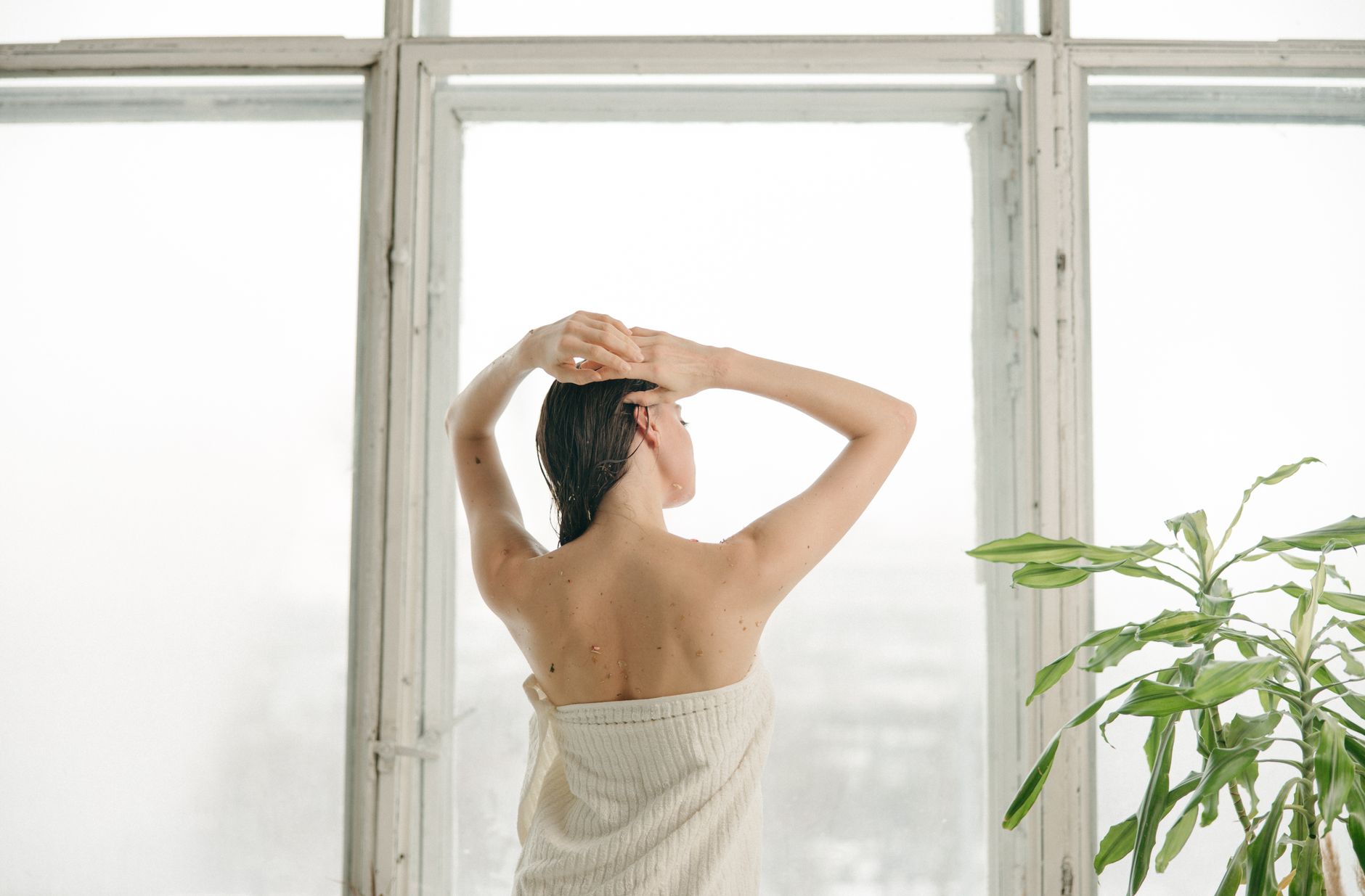 A woman wrapped in a towel stands by a large window, embracing a serene moment.